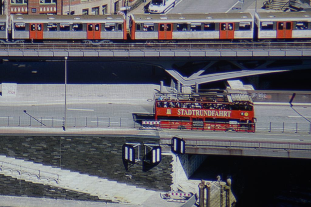 Begeistert bin ich auch von der Projektion meines Hamburg-Panoramas, das ich aus der 21. Etage des Hanseatic Trade Centers geschossen habe. In der 5 % großen Ausschnittsvergrößerung (siehe Foto) ist auffällig, wie präzise der Schriftzug „STADTRUNDFAHRT“ auf dem roten Doppeldecker reproduziert wird. Auch die orangenen U-Bahntüren erscheinen originalgetreu. Der Asphalt ist farbneutral. Hier fallen die Graustufenverfärbungen nicht auf.